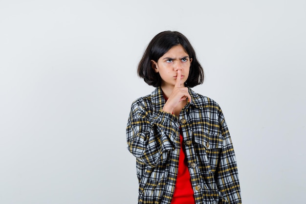 Jong meisje dat stiltegebaar in geruit overhemd en rood t-shirt toont en boos kijkt, vooraanzicht.