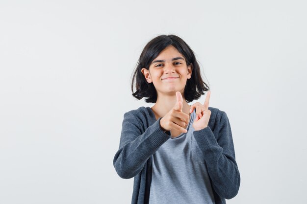 Jong meisje dat tegengestelde richtingen wijst met wijsvingers in lichtgrijs t-shirt en donkergrijze hoodie met ritssluiting en er gelukkig uitziet,