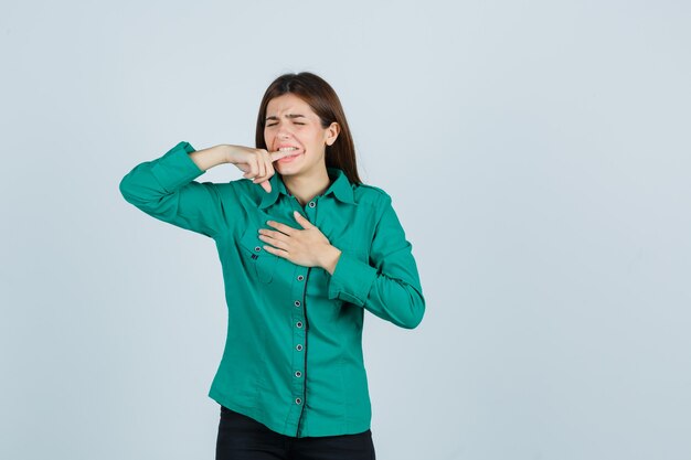 Jong meisje dat wijsvinger bijt, hand over borst in groene blouse, zwarte broek houdt en uitgeput kijkt. vooraanzicht.