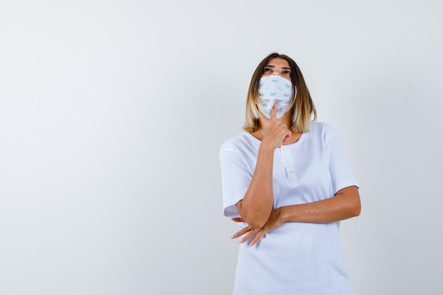 Jong meisje dat wijsvinger onder kin zet, hand onder elleboog in wit t-shirt en masker houdt en peinzend kijkt. vooraanzicht.