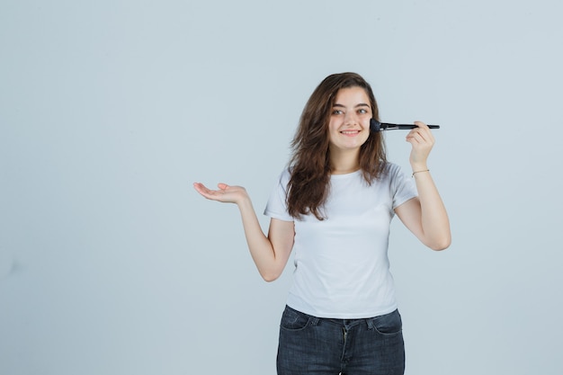 Jong meisje doet make-up met borstel terwijl ze doet alsof ze iets in t-shirt, spijkerbroek laat zien en er vrolijk uitziet. vooraanzicht.