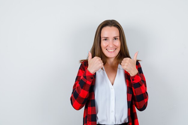 Jong meisje duimen opdagen in geruit hemd, blouse en op zoek vrolijk, vooraanzicht.