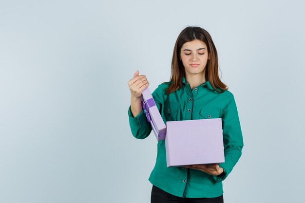 Jong meisje geschenkdoos openen in groene blouse, zwarte broek en gericht kijken. vooraanzicht.