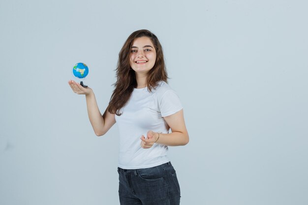 Jong meisje globe houden terwijl kijken naar camera in t-shirt, spijkerbroek en op zoek naar mooi, vooraanzicht.