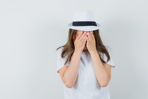 Jong meisje hand in hand op gezicht in wit t-shirt, hoed en op zoek depressief, vooraanzicht.