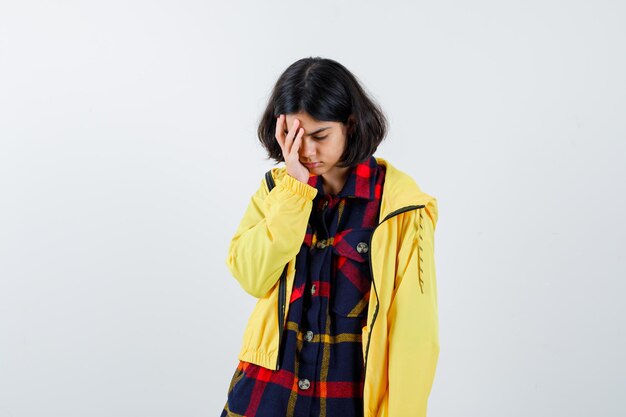 Jong meisje hand op gezicht in geruit overhemd en gele jas en ziet er moe uit, vooraanzicht.