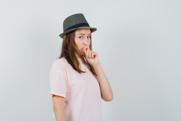 Jong meisje hand op kin in roze t-shirt, hoed en aarzelend op zoek. vooraanzicht.