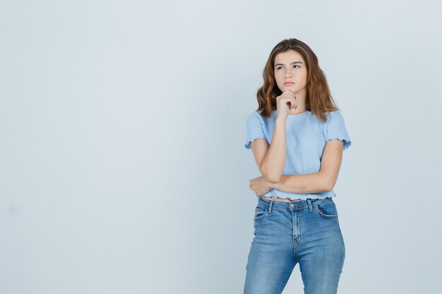 Jong meisje hand op kin in t-shirt, spijkerbroek houden en op zoek doordachte, vooraanzicht.