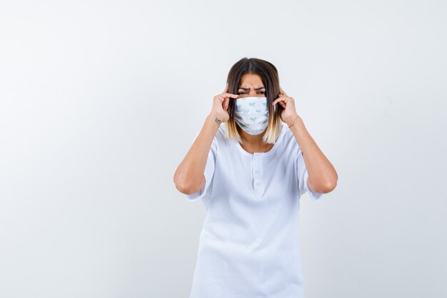 Jong meisje handen op de oren te drukken, grimassen in wit t-shirt, masker en op zoek gekwelde, vooraanzicht.