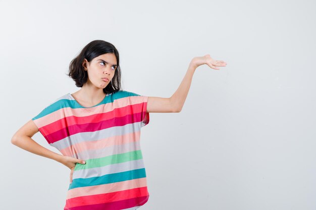 Jong meisje houdt de hand op de taille terwijl ze de hand uitrekt terwijl ze iets vasthoudt in een kleurrijk gestreept t-shirt en er geïrriteerd uitziet. vooraanzicht.