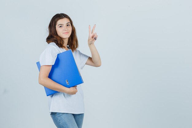 Jong meisje in de witte map van de t-shirtholding, die overwinningsgebaar toont en er zelfverzekerd uitziet, vooraanzicht.