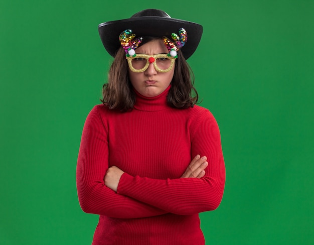 Jong meisje in een rode trui met grappige bril en zwarte hoed camera kijken ontevreden met gekruiste armen staande over groene achtergrond