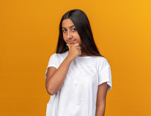 Jong meisje in een wit t-shirt dat naar de camera kijkt met een peinzende uitdrukking op het gezicht met de hand op haar kin die over oranje staat