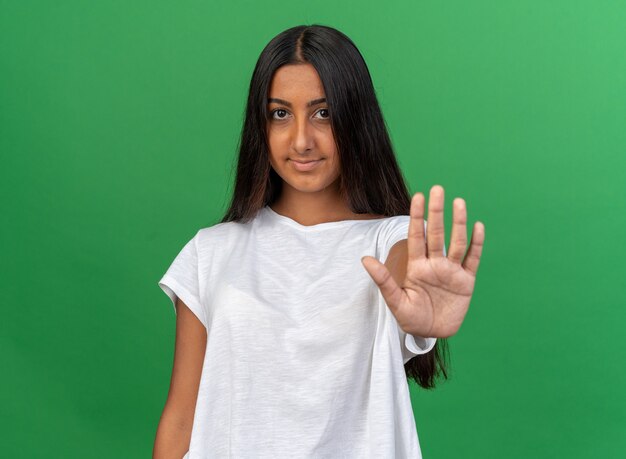 Jong meisje in een wit t-shirt dat naar de camera kijkt met een zelfverzekerde glimlach op het gezicht en een stopgebaar maakt met de hand