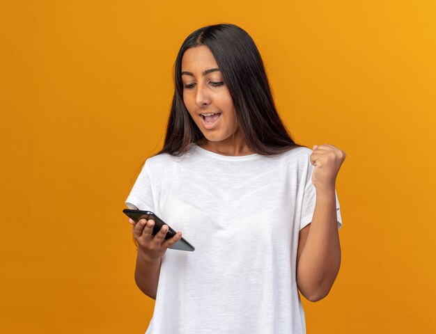 Jong meisje in een wit t-shirt met een smartphone die ernaar kijkt en de vuist balt, blij en opgewonden
