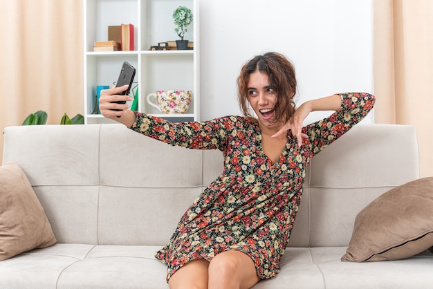 Jong meisje in gebloemde jurk doet selfie met smartphone gelukkig en positief glimlachend vrolijk zittend op een bank in lichte woonkamer