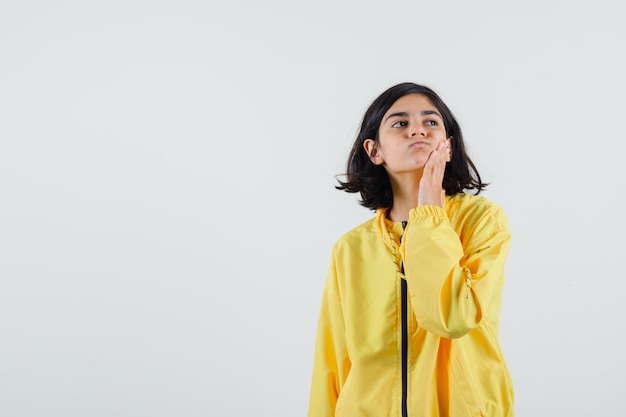 Jong meisje in geel bomberjack staande in denken pose, hand op de wang zetten en peinzend kijken