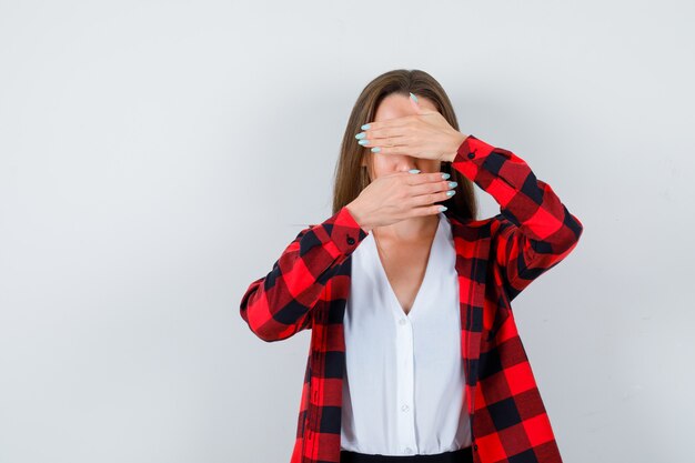 Jong meisje in geruit hemd, blouse die gezicht bedekt met handpalmen en er mysterieus uitziet, vooraanzicht.