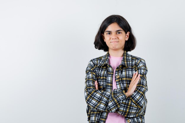 Jong meisje in geruit overhemd en roze t-shirt staande armen gekruist en zelfverzekerd, vooraanzicht.