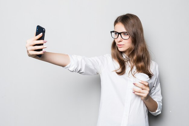 Jong meisje in glazen gekleed in strikte kantoor wit t-shirt staat voor witte muur en houdt haar telefoon in handen