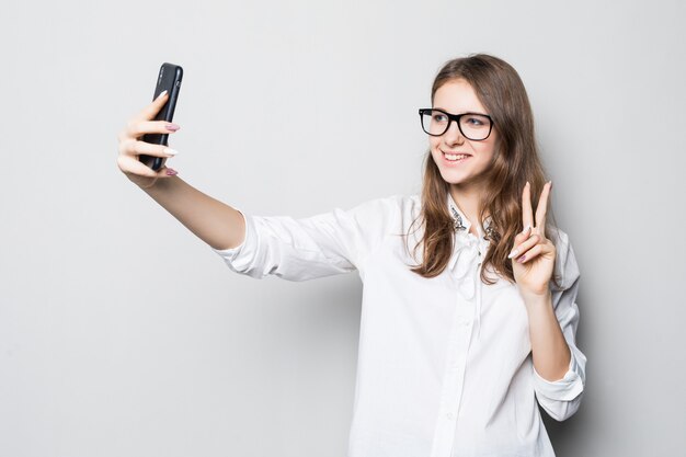 Jong meisje in glazen gekleed in strikte kantoor wit t-shirt staat voor witte muur en houdt haar telefoon in handen