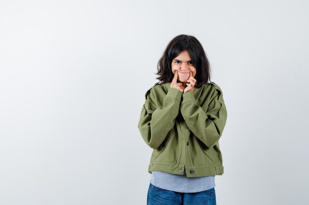 Jong meisje in grijze trui, kaki jasje, jeansbroek met wijsvingers in de buurt van mond, dwingt een glimlach en ziet er schattig uit, vooraanzicht.