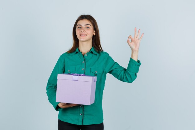 Jong meisje in groene blouse, zwarte broek die giftdoos houdt, ok teken toont en vrolijk, vooraanzicht kijkt.