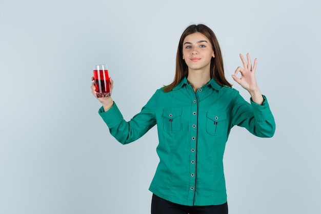 Jong meisje in groene blouse, zwarte broek die glas rode vloeistof houdt, ok teken toont en gelukkig, vooraanzicht kijkt.