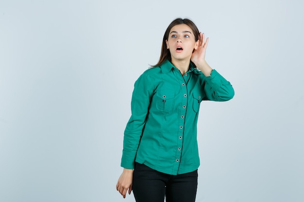 Jong meisje in groene blouse, zwarte broek die hand dichtbij oor houdt om iets te horen en geconcentreerd, vooraanzicht kijkt.