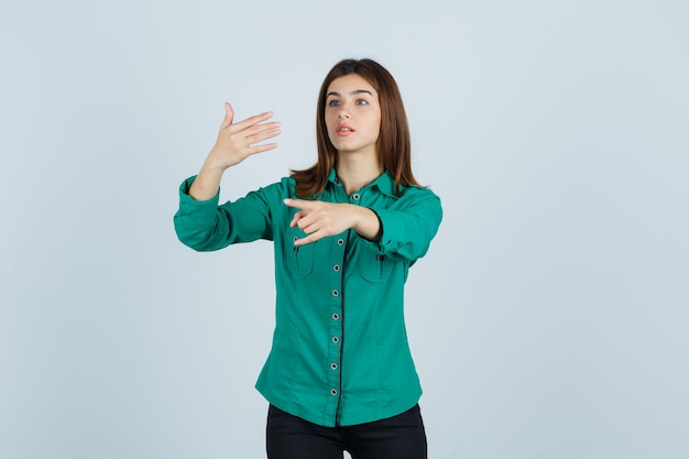 Jong meisje in groene blouse, zwarte broek die hand uitrekt als iets denkbeeldigs vasthoudt, rock n roll-gebaar toont en gefocust, vooraanzicht kijkt.