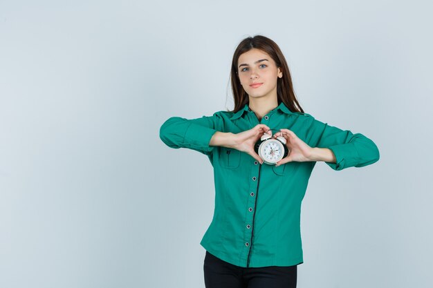 Jong meisje in groene blouse, zwarte broek die klok in beide handen houdt en gelukkig, vooraanzicht kijkt.