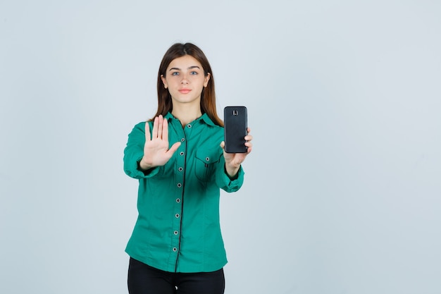 Jong meisje in groene blouse, zwarte broek die telefoon in één hand houdt, stopbord toont en ernstig, vooraanzicht kijkt.