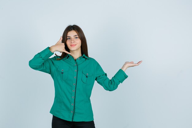 Jong meisje in groene blouse, zwarte broek die telefoongebaar toont, palm opzij spreidt en optimistisch, vooraanzicht kijkt.