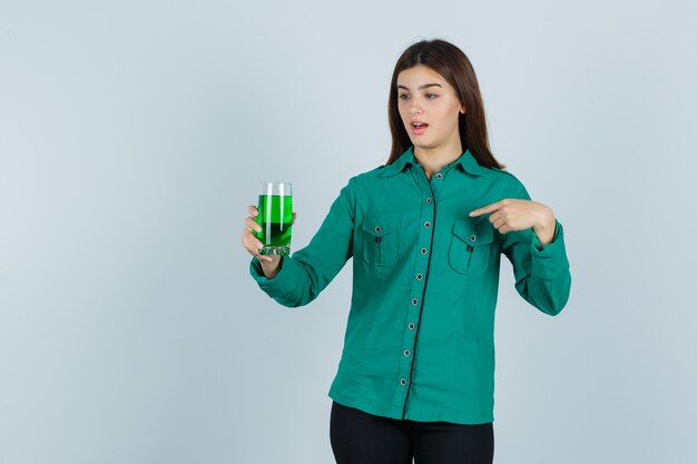 Jong meisje in groene blouse, zwarte broek met een glas groene vloeistof, erop wijzend met wijsvinger en geschokt, vooraanzicht op zoek.