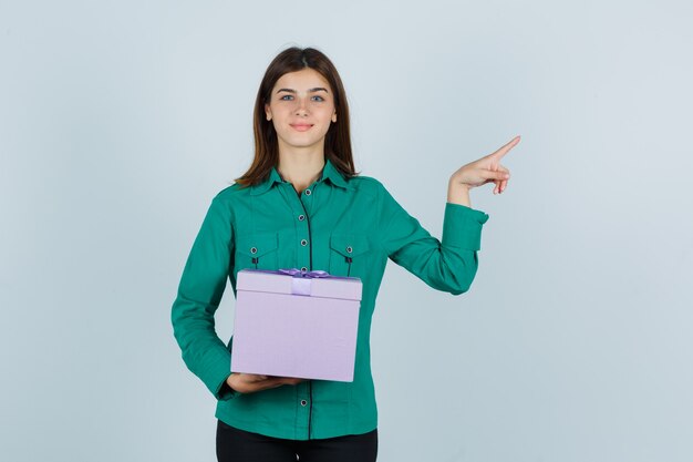 Jong meisje in groene blouse, zwarte broek met geschenkdoos, naar rechts wijzend met wijsvinger en op zoek gelukkig, vooraanzicht.