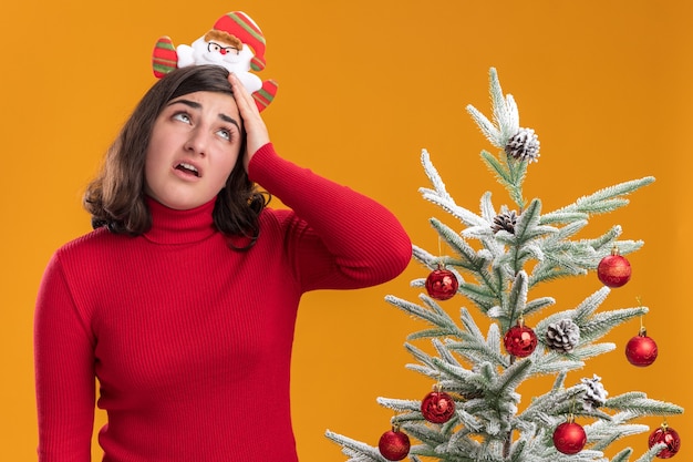 Jong meisje in Kerstmissweater die grappige hoofdband draagt die ogen moe en verveeld oprolt naast een kerstboom over oranje muur