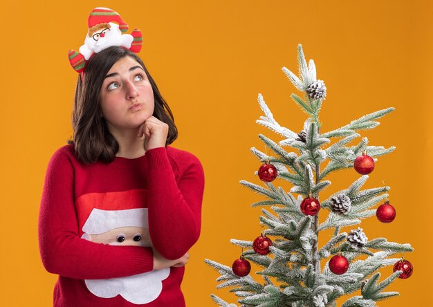 Jong meisje in Kerstmissweater die grappige hoofdband draagt die verbaasd status naast een kerstboom over oranje muur kijkt