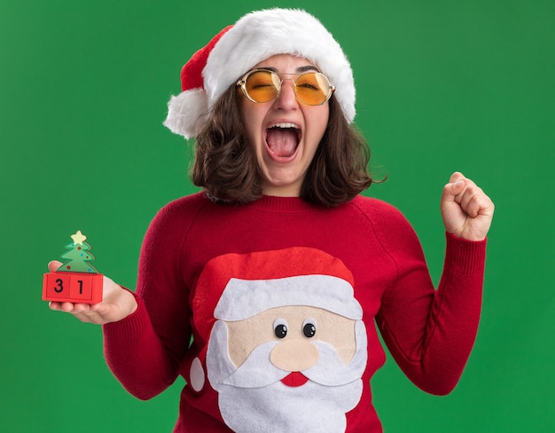 Gratis foto jong meisje in kerstmissweater die kerstmuts en bril draagt ?? die stuk speelgoed kubussen met nieuwjaarsdatum houdt gek, blij en opgewonden gebalde vuist die zich over groene muur bevindt