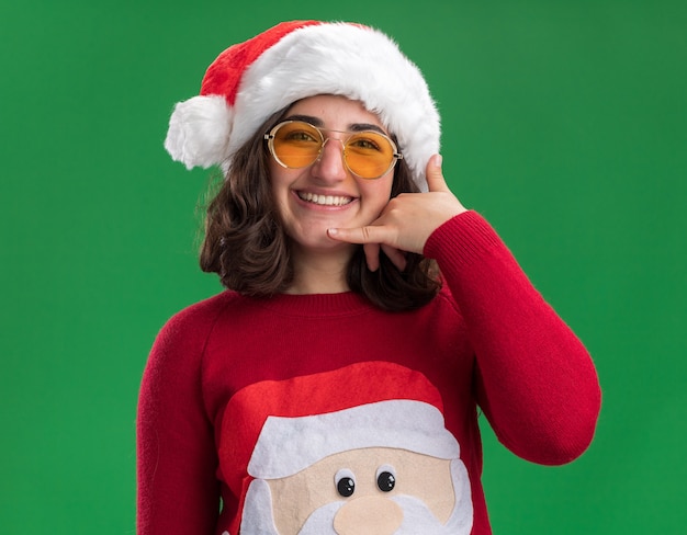 Jong meisje in Kerstmissweater die santahoed en glazen dragen die bellen me gebaar glimlachen die vrolijk zich over groene muur bevinden