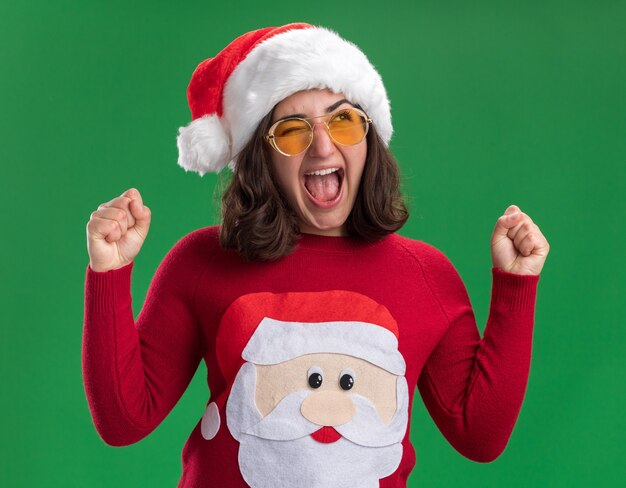 Jong meisje in Kerstmissweater die santahoed en glazen dragen die opgewonden en gekke gelukkige gebalde vuisten schreeuwen die zich over groene muur bevinden
