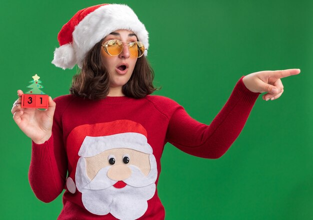 Jong meisje in Kerstmissweater die santahoed en glazen dragen die stuk speelgoed kubussen met nieuwjaarsdatum houden