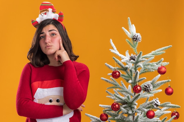 Jong meisje in kersttrui met een grappige hoofdband die opzij kijkt en verbaasd naast een kerstboom staat over oranje achtergrond