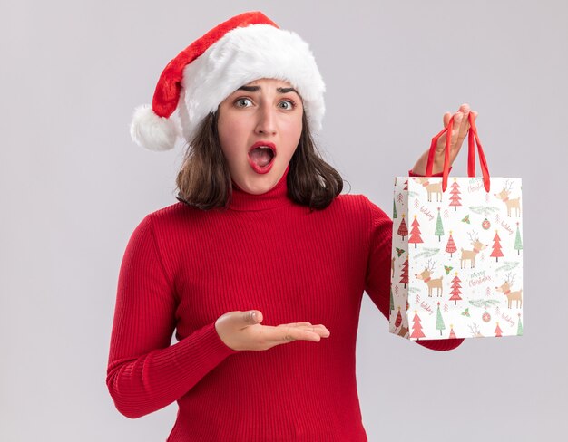 Jong meisje in rode trui en kerstmuts bedrijf kleurrijke papieren zak met kerstcadeaus op zoek verrast presenteren met arm van haar hand staande op witte achtergrond