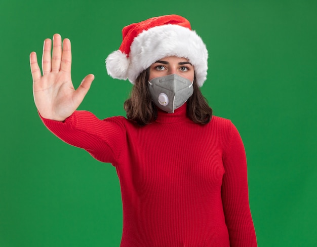 Gratis foto jong meisje in rode trui en kerstmuts die gezichts beschermend masker met ernstig gezicht dragen dat stopgebaar met hand maakt die zich over groene muur bevindt