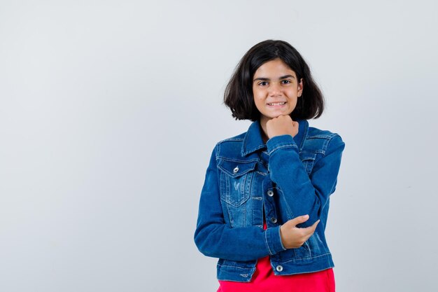 Jong meisje in rood t-shirt en spijkerjasje leunend met kin bij de hand, glimlachend en gelukkig kijkend