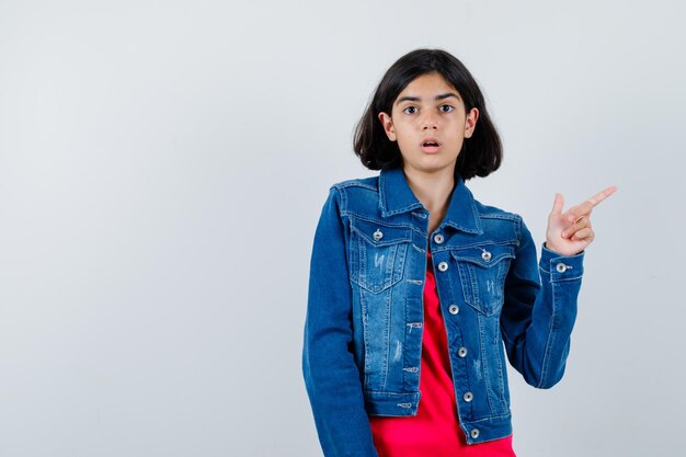 Jong meisje in rood t-shirt en spijkerjasje naar rechts wijzend met wijsvinger en verbaasd kijken, vooraanzicht.