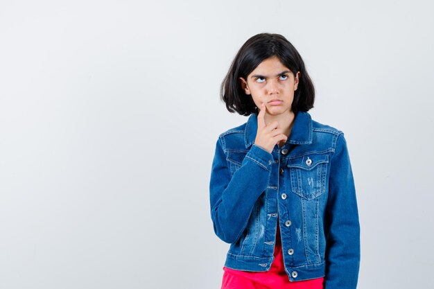 Jong meisje in rood t-shirt en spijkerjasje staat in denkende pose en kijkt peinzend