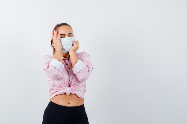 Jong meisje in roze blouse, zwarte broek, masker met een hand op de tempel, een andere hand op de kaak, de ogen gesloten houden en er moe uitziet, vooraanzicht.