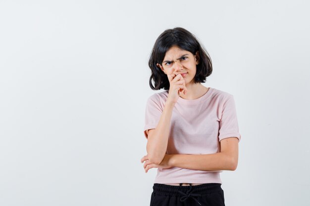 Jong meisje in roze t-shirt en zwarte broek die zich in het denken stelt en hand op kin zet en peinzend kijkt