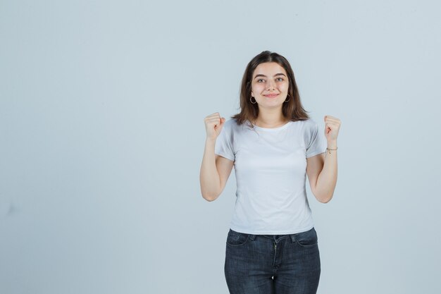 Jong meisje in t-shirt, jeans die winnaargebaar toont en gelukkig, vooraanzicht kijkt.